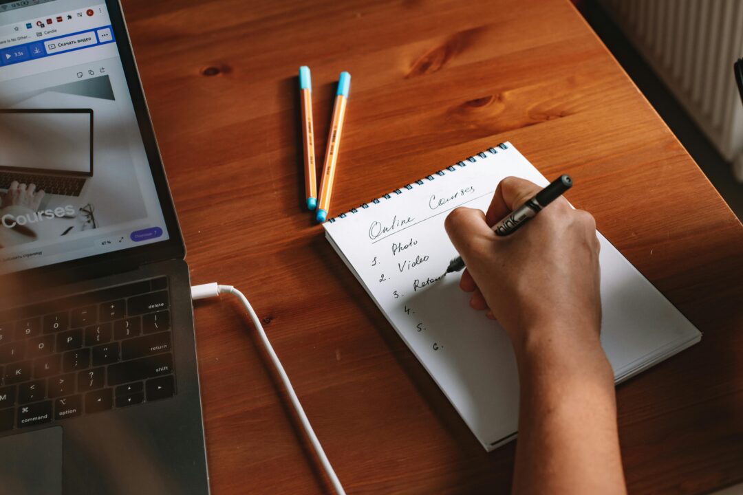 pexels-photo-5912284-5912284-1 Photo of a person writing a list of online courses on a notepad next to a laptop.